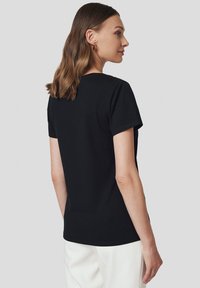 Czarny t-shirt z krótkim rękawem, miękka tkanina, okrągły dekolt, dopasowany krój, minimalistyczny design, noszony z białymi spodniami, widok z tyłu.