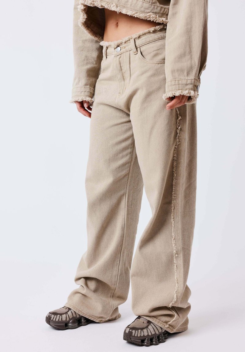 Personne portant un jean beige à jambes larges avec des coutures latérales effilochées et une veste courte assortie, associée à des baskets foncées à motifs.