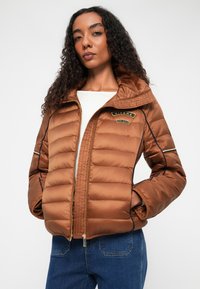 Jachetă puff de culoare brown cu quilt-ing orizontal, închidere cu fermoar și glugă. Prezintă un patch cu logo și detalii contrastante negre pe mâneci.