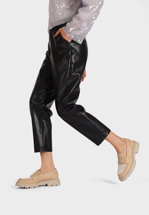 Mocassini beige con una finitura testurizzata e dettagli con logo, abbinati a pantaloni neri in ecopelle slim. Elementi riflettenti sulle scarpe.