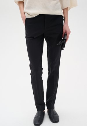 Černé tailored kalhoty s hladkou strukturou, vybavené bočními kapsami a slim-fit střihem, doplněné černými nazouvacími botami.