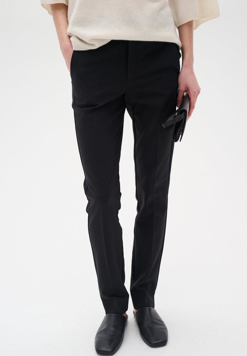 Černé tailored kalhoty s hladkou strukturou, vybavené bočními kapsami a slim-fit střihem, doplněné černými nazouvacími botami.