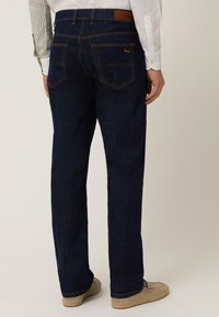Temno modri denim hlače s ravnimi nogavicami, vidnimi zadnjimi žepi, oranžnim šivanjem in rjavim usnjenim podstavkom na pasu.