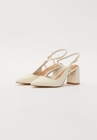 Beige kiilto-kengät, joissa on terävä kärki, paksu lohkokorko ja säädettävä nilkkahihna; valmistettu sileästä nahasta ja metallisista yksityiskohdista.