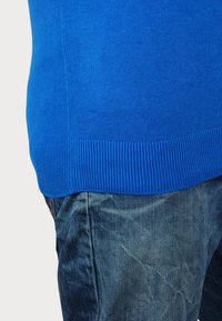 Modra pletena jopica z rebrastim robom, v kombinaciji z teksturiranimi kavbojkami, ki prikazujejo zbledela vzorca in sproščen kroj.