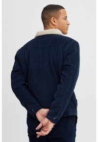 Chaqueta de corduroy azul marino con un collar de borreguito crema, puños abotonados y un detalle en el yugo trasero, diseñada con un ajuste relajado.
