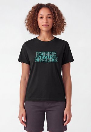 Jonge vrouw met krullend haar, die een zwart T-shirt met turquoise tekst "BONNE CHANCE" en donkere korte broek draagt tegen een effen achtergrond.