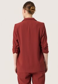 Sacou bordeaux cu o textură netedă, croială ajustată și mâneci pliate. Se remarcă prin reverele tradiționale și un design curat, simplu pe spate.