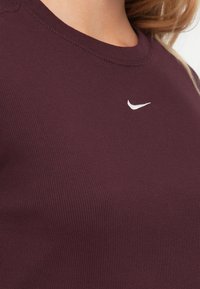 Burgundi spalvos raukšlių audinio marškinėliai su apvalia iškirpte ir maža balta Nike logo, esanti krūtinės centre. Lygus paviršius.