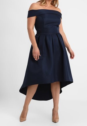 Mujer vistiendo un vestido azul marino con hombros descubiertos, bajo asimétrico y falda plisada, combinado con tacones puntiagudos en color nude.