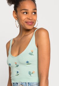 Top curto verde menta com decote em V, apresentando bordados florais em laranja, azul e verde. Tecido tricotado e detalhe de bainha canelada.