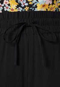 Pantalon en tissu noir avec une taille élastique et un cordon noué, associé à un haut fleuri avec des fleurs jaunes, bleues et rouges.