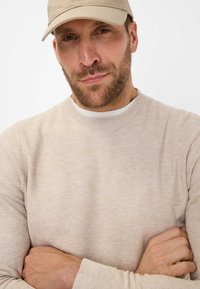 Helles beige Strickpullover mit strukturierter Oberfläche und Rundhalsausschnitt, kombiniert mit einer hellbeigen Baseballkappe. Arme vor der Brust verschränkt.