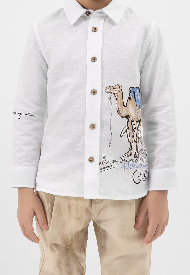 Enfant portant une chemise blanche boutonnée avec une illustration de chameau et un pantalon beige, debout devant un fond uni.