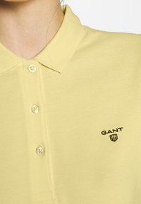 Polo shirt jaune clair avec un tissu texturé, patte de boutonnage à trois boutons et logo "GANT" noir brodé sur la poitrine gauche.