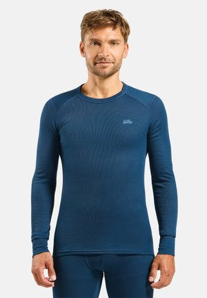 Mann trägt enganliegendes blaues Thermooberteil mit langen Ärmeln und passende Unterteile, steht vor einem schlichten hellen Hintergrund.