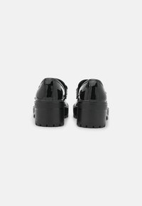 Chaussures en cuir verni noir avec un talon épais, présentant une finition lisse et une semelle striée pour une meilleure adhérence. La vue arrière montre des bords arrondis.