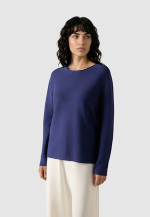 Kvinde med bølget sort hår iført en langærmet marineblå sweater og løstsiddende hvide bukser, der kigger til siden mod en ensfarvet baggrund.