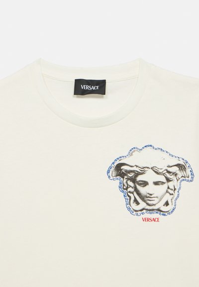 Μπεζ βαμβακερό t-shirt με κεντρικό σχέδιο το πρόσωπο της Μέδουσας, περιγραμματισμένο με μπλε και με μοτίβο ελληνικού κλειδιού, καθώς και κόκκινο κείμενο "VERSACE".