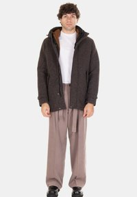 Uomo in piedi che indossa una giacca marrone con cappuccio sopra una camicia bianca, pantaloni beige larghi e scarpe nere su uno sfondo bianco uniforme.