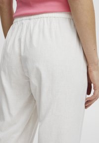 Pantalons en lin blanc avec une taille élastique, présentant une texture lisse et une coupe ample, partiellement visibles de côté.