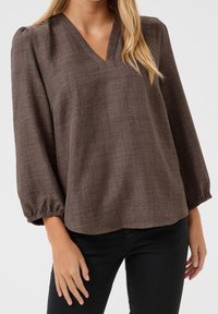 Blusa marrón texturizada con escote en V y mangas abullonadas, hecha de un tejido ligero. Corte casual, combinada con pantalones negros.