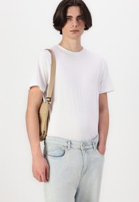 Sac bandoulière léger beige avec une sangle réglable, porté sur un t-shirt blanc uni et associé à un jean en denim bleu clair.