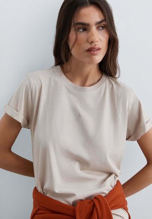 Mujer de cabello oscuro que lleva una camiseta de manga remangada de color beige claro y un suéter color óxido atado alrededor de su cintura sobre un fondo liso.