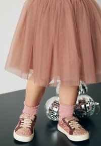 Rosa Tüllrock mit geschichteter Gestaltung. Funkelnde rosa und braune Sneakers mit Klettverschluss, kombiniert mit gerippten rosa Socken.