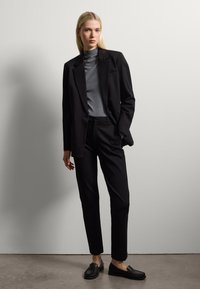 Zwarte op maat gemaakte pak met een single-button jasje, grijze turtleneck en zwarte instapschoenen. Gladde stof, slim fit, minimalistisch ontwerp.