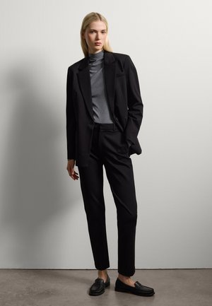 Traje negro a medida con chaqueta de un solo botón, suéter de cuello alto gris y mocasines negros. Tela suave, corte ajustado, diseño minimalista.