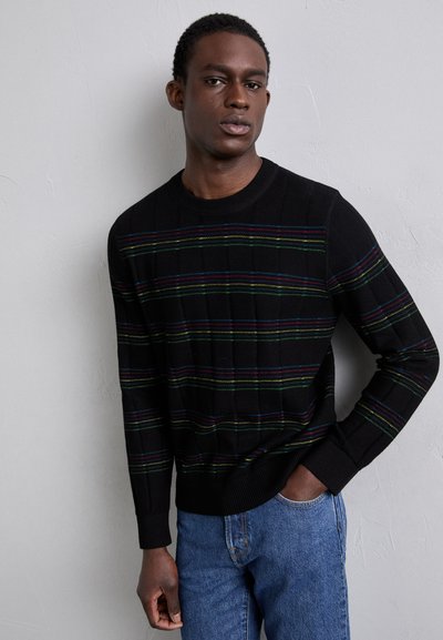 Pull noir en tricot avec de vives rayures horizontales vertes, bleues et rouges. Il présente un col rond et des poignets côtelés. Porté avec un jean bleu.