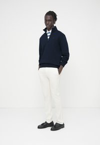 Μπλε ναυτικό πουλόβερ με φερμουάρ, λευκό πουκάμισο με μπλε ρίγες στο γιακά, κρεμ παντελόνι και μαύρα μοκασίνια. Απλές γραμμές και αντιθετικά χρώματα.