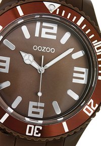 Montre en caoutchouc marron avec un cadran marron, des marqueurs d'heure blancs et des aiguilles argentées. Elle dispose d'une lunette tournante marron avec des chiffres et des accents blancs.