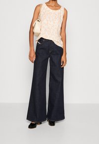 Haut sans manches blanc à motif floral associé à un jean large bleu foncé. Le modèle tient un sac à main beige et porte des chaussures à talons blocs noires.