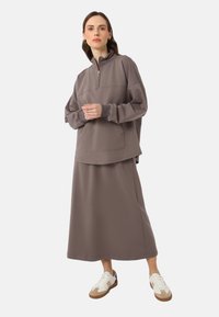 Brauner Fleece-Pullover mit Viertelreißverschluss, seitlichen Taschen und passendem, knöchellangem Rock. Getragen mit weißen und braunen Sneakers. Sanfte Textur.