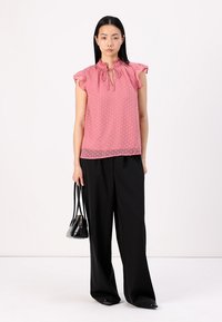 Blusa rosa a pois con maniche a volant e colletto con fiocco. Abbinata a pantaloni neri larghi e una piccola borsa nera. Tessuto morbido.