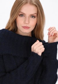 Tengerészkék kötött pulóver bordás textúrával, széles gallérral és dús ujjakkal. Puha anyag laza szabással és finom részletezéssel.