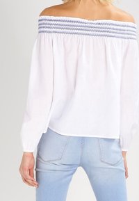 Haut blanc épaules dénudées avec détails bleus froncés, manches longues, coupe ample, associé à un jean bleu clair. Texture de tissu douce.