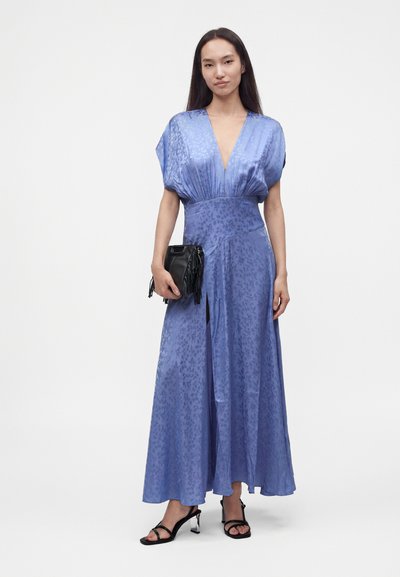 Femme portant une longue robe bleue à motifs avec un décolleté en V profond, tenant une pochette noire à franges et portant des sandales à talons noires.