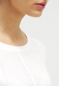 Hvid, kortærmet bluse med rund halsudskæring og knaplukning. Stoffet fremstår let og glat med en ren finish.