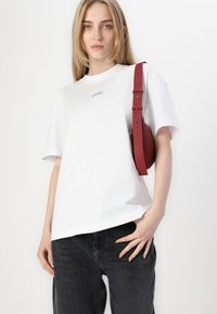 Hvit bomulls t-skjorte med rund hals og korte ermer. Har en liten merket logo på brystet. Sammen med svarte jeans og en rød bag.