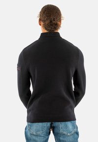 Pullover nero a coste con colletto alto, vestibilità slim e sottile accento rosso sulla manica sinistra, abbinato a jeans azzurri chiari.