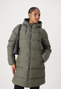 Manteau matelassé vert olive, extérieur en nylon, design piqué, fermeture éclair à l'avant, capuche ajustable, deux poches latérales, associé à un sac à main noir.