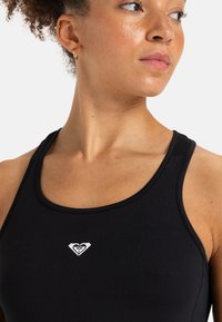 Musta urheilullinen t-paita pyöreällä kaula-aukolla, jossa keskellä on pieni valkoinen logo. Sujuva kangas ja vartalonmyötäinen muotoilu.