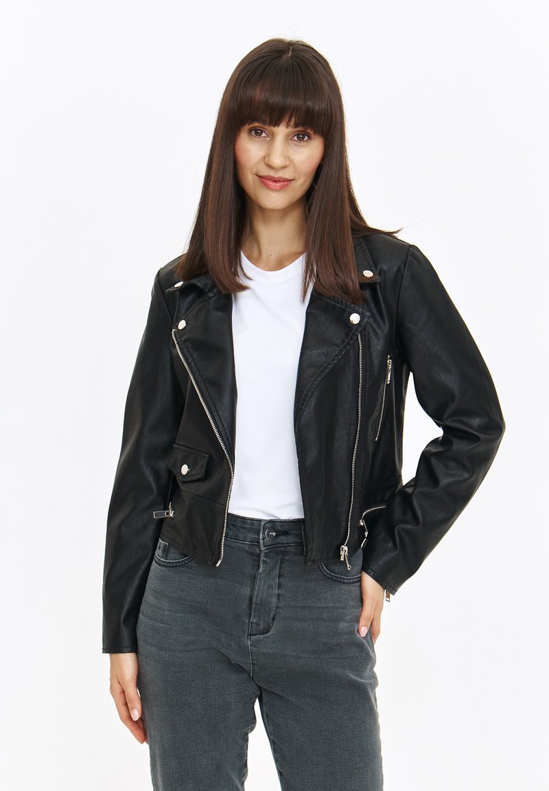 Czarna skórzana kurtka motocyklowa z srebrnymi akcentami, zamykanymi kieszeniami i dopasowanym krojem. Noszona na białej koszuli i zestawiona z szarymi dżinsami.
