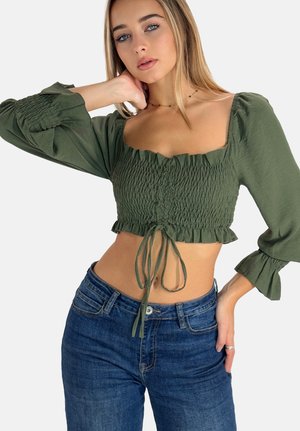 Jonge vrouw met lang blond haar draagt een geplooide olijfgroene cropped top met lange mouwen en een high-waisted blauwe spijkerbroek tegen een witte achtergrond.