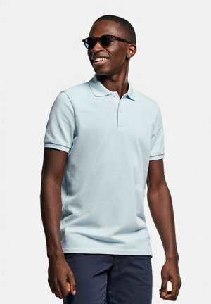 Polo bleu ciel en tissu texturé, avec un col classique et des manches courtes. Porté avec un short foncé et des lunettes de soleil.