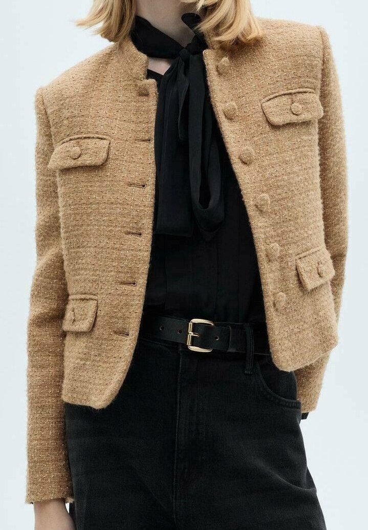 Femme portant une veste texturée couleur chameau avec des poches boutonnées, un chemisier noir avec un nœud au col, un pantalon noir et une ceinture noire avec une boucle dorée.