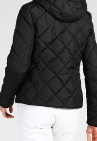 Veste matelassée noire avec capuche, motif losange, poignets élastiques et silhouette cintrée, avec un extérieur lisse et brillant.